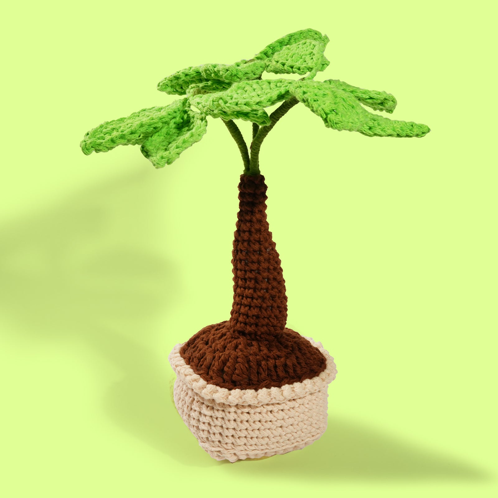 A handcrafted crochet palm tree with green leaves and a brown trunk in a beige basket, set against a yellow background.