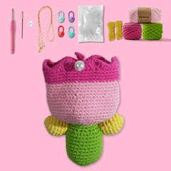 A collection of crafting materials including yarn in pink, green, and yellow, crochet hooks, and other accessories to create a tulip-shaped bag, displayed on a pink background.